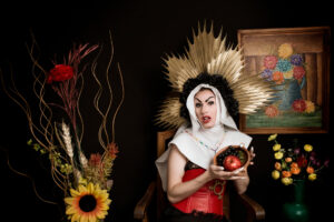 Imagen de la nota "La santa víscera", introducción a Revista La Cascotiada #2. Fotografia de una drag queen vestida al estilo de una monja con un gran tocado en la cabeza y sostiendo una vasija con una manzana. Al lado se observan dos floreros con flores secas y de fondo un cuadro de una naturaleza muerta.